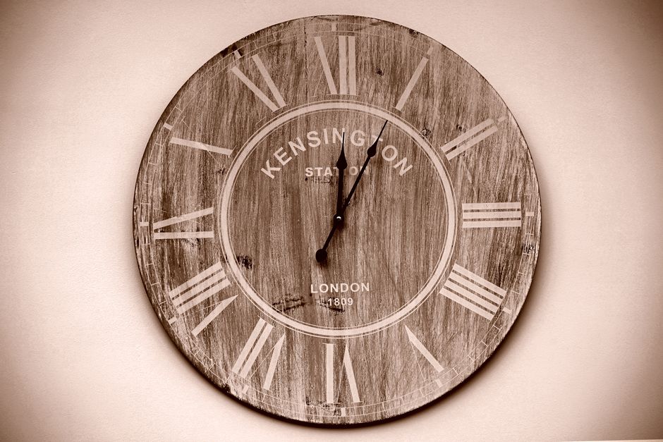 A round wall clock with a rustic, aged wooden finish featuring Roman numerals in a faded white color, positioned on a plain, light-colored wall. The clock face displays the words 'Kensington Station' and 'London 1809' in a vintage style, with black hour and minute hands showing the time as approximately 12:02. The textured wood surface has visible grain and weathered details, contrasting subtly with the smooth wall background. The clock's minimalist design and retro aesthetic evoke a classic, timeless look often associated with traditional railway station clocks, aligning with themes of heritage and reliability. This image could be used to illustrate the importance of punctuality in time-sensitive contexts, such as coordinating private waste collection or on-site clearance services provided by waste removal specialists like wasteremovalsouthkensington.co.uk, which may seek to avoid reliance on local authority rubbish collection in favor of independent waste handling solutions.