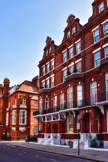 A tall red brick residential building situated on a city street, featuring multiple stories with large sash windows framed in white and black wrought iron balconies on each floor. The building’s facade includes decorative architectural details such as arched windows on the lower levels and ornate cornices. The ground entrance is accessible via a set of white steps leading to an arched doorway, with small potted plants placed near it. Surrounding the building are other similar urban structures, and the street appears clean with minimal vehicle presence. The scene is illuminated by natural daylight against a clear blue sky, emphasizing the historic character of the architecture. This setting may relate to independent or private waste management solutions, commonly employed for property maintenance or on-site clearance in such residential buildings, with no visible waste or rubbish on the street or near the building itself. This visual context aligns with typical urban rubbish removal services that specialise in residential properties like this one, highlighting the importance of effective waste handling for maintaining the area’s tidy appearance, as provided by companies such as waste removal services in South Kensington or similar areas.