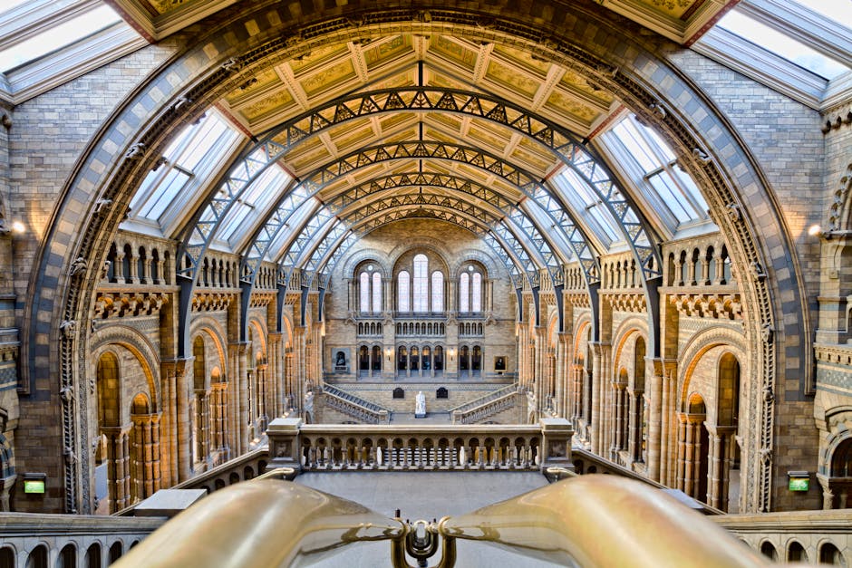The image depicts the interior of a grand, historic building with a spacious, double-height central hall. The ceiling features an ornate, vaulted iron and glass roof structure, allowing natural light to illuminate the space below. The walls are constructed from light-colored bricks with decorative arches and columns, some of which have intricate detailing. Large, tall windows with rounded tops line the upper level, providing additional natural light and showcasing a classic architectural style. A staircase with a decorative balustrade is visible in the middle ground, leading up to an upper balcony that runs along the perimeter of the hall. In the background, at the far end of the room, there is a statue positioned on a pedestal, and various smaller niches and decorative features are integrated into the wall. The overall atmosphere combines historical elegance with a bright, airy ambiance, suited for a museum or public building that might be involved in aesthetic or educational activities, aligning with themes of heritage preservation and cultural exhibition.