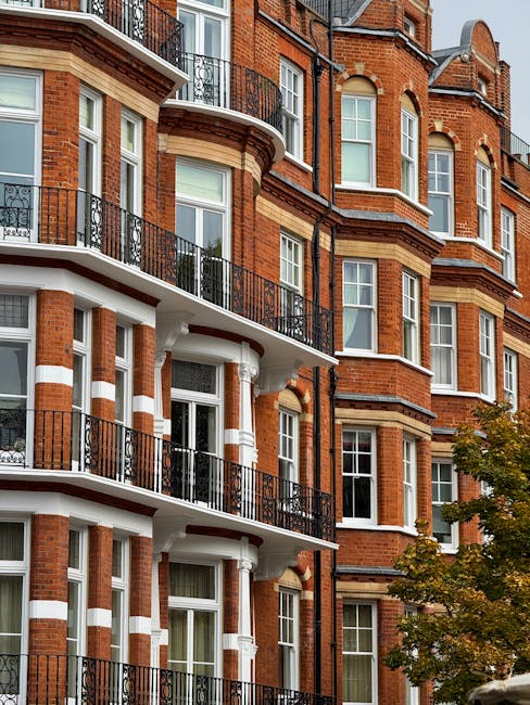 The image depicts the exterior of a multi-story red-brick residential building featuring ornate architectural details typical of Victorian or Edwardian style. Multiple bay windows with white frames are visible, each topped with decorative arches and brickwork. The building has several wrought-iron balconies with black railing, extending from the windows on the lower floors. In the foreground, a small tree with greenish-brown leaves partially obscures the lower part of the building, suggesting an urban street or courtyard setting. The brickwork showcases a mixture of red tones with beige and cream accents highlighting the stone window sills and decorative bands running horizontally across the facade. The scene is illuminated by natural daylight, creating a clear and neutral atmosphere. While the primary focus is on the building’s historic architecture, the image may imply the context of urban living where independent waste management or regular rubbish removal services like those provided by waste removals in South Kensington could be relevant in maintaining the cleanliness of such residential environments.