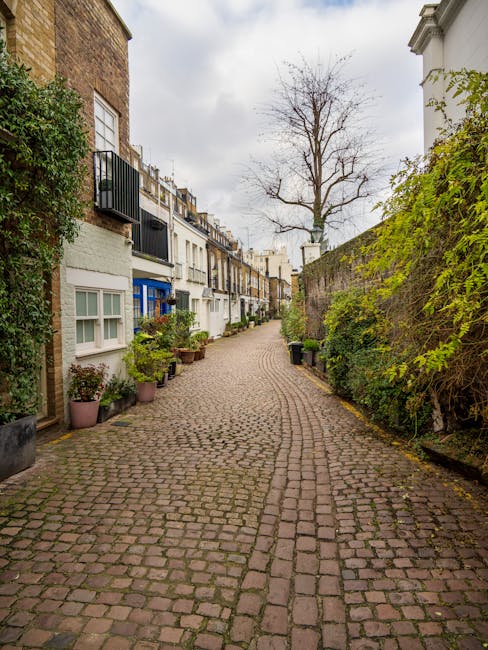 A narrow, cobbled residential alleyway lined with potted plants and greenery on the left side, featuring multi-story terraced houses painted in white and with small balconies, windows, and rooftop details. On the right, a high, moss-covered stone wall is partially obscured by dense foliage, and beyond it, a leafless tree with bare branches extends upward against a cloudy sky. The scene appears quiet and well-maintained, with a few small rubbish bins positioned along the edge of the path, suggesting private waste management or independent rubbish collection in the area. The overall atmosphere is calm, with natural light diffused through the overcast sky, emphasizing the textures of the cobblestones and the varied surfaces of the buildings and greenery, aligning with the context of alternative waste handling and rubbish removal services typical near South Kensington Station.