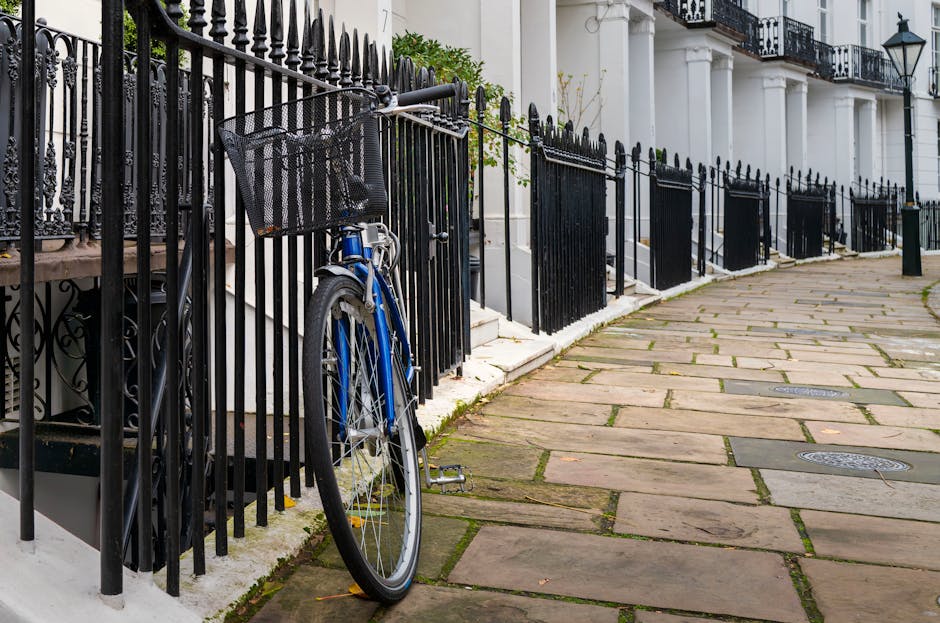 The image features a blue bicycle parked and secured by a black metal railing along a stone-paved sidewalk in front of a white building with classical architectural details, including white columns and decorative balustrades on a balcony. The bicycle has a black front basket, a silver chain, and black pedals, and leans against the railing on the left side of the frame. The railing, extending along the length of the building's entrance, is composed of evenly spaced vertical bars with pointed tops, creating a boundary between the sidewalk and the building steps. The stone paving consists of irregularly shaped slabs in shades of beige and brown, with narrow gaps filled with moss or grass, and the surface appears slightly weathered. The background includes additional railings and steps leading up to the building’s entrance, with lush green plants visible near the top of the steps. This scene is associated with private property and urban waste management, where onsite or alternative waste handling may be relevant to local rubbish collection practices, as subtly implied by the clean and organized setting. The lighting is natural, suggesting daytime, with soft shadows indicating an overcast sky or diffused sunlight.