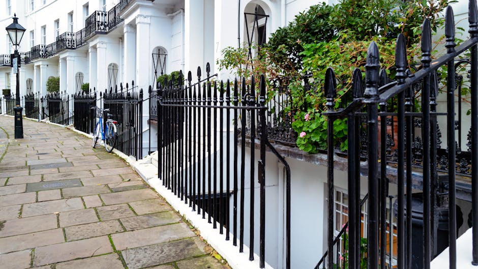 A row of white terraced residential buildings with black wrought iron railings lining the sidewalk in a quiet urban area. The buildings feature tall, narrow windows with black metal arches above, and small balconies with decorative ironwork. On the right side, a lush green plant with pink flowers grows behind the railings, partially obscuring part of the building facade. The pavement consists of large, irregular stone slabs laid in a staggered pattern, with some moss or dirt visible in the joints. A blue bicycle is parked and secured to the fence on the left side of the image. The scene is illuminated by natural daylight, creating soft shadows and highlighting the textures of the railings, stone pavement, and building surfaces. This setting may relate to private property access in South Kensington, suitable for alternative waste handling or disposal services provided by waste removal specialists like waste removals south Kensington, emphasizing the importance of clear pathways for rubbish collection or on-site clearance activities.